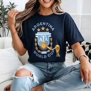 以下が含まれることがあります： アルゼンチン2026年ワールドカップのロゴが入ったネイビーブルーのTシャツ。デザインには、アルゼンチンサッカー協会のエンブレム、星、金色のトロフィーが含まれています。Tシャツはブルージーンズと合わせて着用されています。