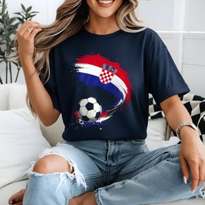 May include: Navy blue t-shirt featuring a Croatian flag design with a soccer ball. The design incorporates red, white, and blue brushstrokes and the Croatian coat of arms. The shirt is a casual wear item.