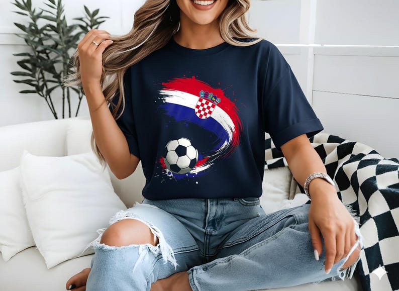 May include: Navy blue t-shirt featuring a Croatian flag design with a soccer ball. The design incorporates red, white, and blue brushstrokes and the Croatian coat of arms. The shirt is a casual wear item.