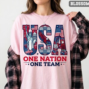 May include: Pink t-shirt with a patriotic USA design. The word "USA" is filled with American flag imagery, the Statue of Liberty, and an eagle. Below, the text reads "ONE NATION ONE TEAM" in red and blue.