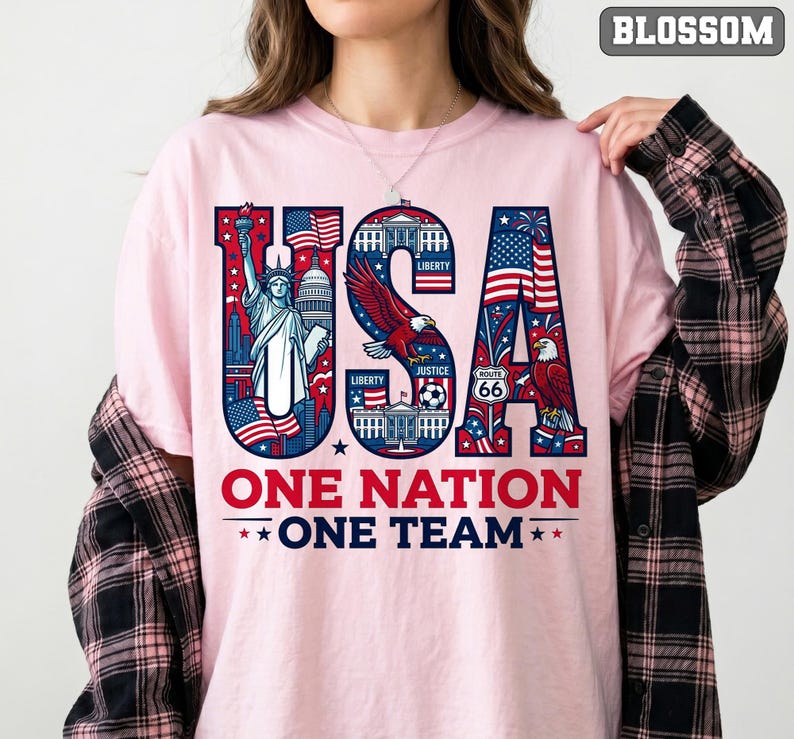 May include: Pink t-shirt with a patriotic USA design. The word "USA" is filled with American flag imagery, the Statue of Liberty, and an eagle. Below, the text reads "ONE NATION ONE TEAM" in red and blue.