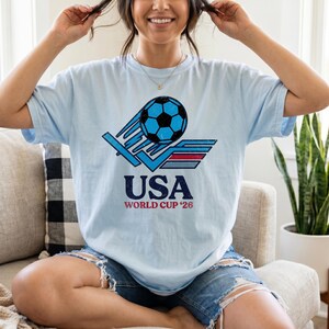 Puede incluir: Camiseta azul claro con un gr&aacute;fico de bal&oacute;n de f&uacute;tbol en azul y negro, y el texto "USA WORLD CUP '26". El dise&ntilde;o tiene un aspecto vintage y desgastado.
