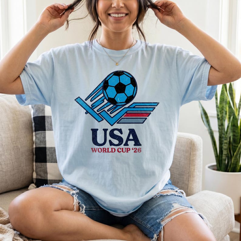 Puede incluir: Camiseta azul claro con un gr&aacute;fico de bal&oacute;n de f&uacute;tbol en azul y negro, y el texto "USA WORLD CUP '26". El dise&ntilde;o tiene un aspecto vintage y desgastado.