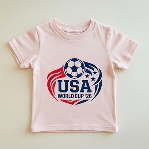 May include: A light pink t-shirt featuring a soccer ball graphic with the text "USA WORLD CUP '26" in navy blue. The design includes red and blue accents with stars, perfect for soccer fans.