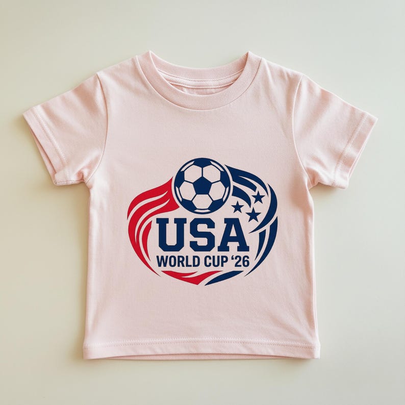 May include: A light pink t-shirt featuring a soccer ball graphic with the text "USA WORLD CUP '26" in navy blue. The design includes red and blue accents with stars, perfect for soccer fans.
