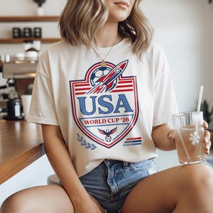 Peut inclure: T-shirt cr&egrave;me avec un graphisme USA World Cup '26. Le design pr&eacute;sente un ballon de football, une fus&eacute;e et les couleurs du drapeau am&eacute;ricain. Le t-shirt est associ&eacute; &agrave; un short en jean.