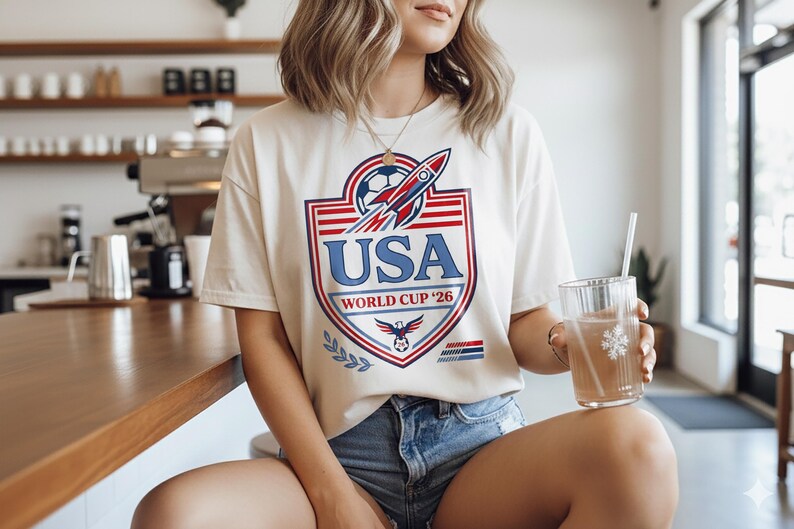 Peut inclure: T-shirt cr&egrave;me avec un graphisme USA World Cup '26. Le design pr&eacute;sente un ballon de football, une fus&eacute;e et les couleurs du drapeau am&eacute;ricain. Le t-shirt est associ&eacute; &agrave; un short en jean.