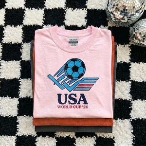 Puede incluir: Una pila de tres camisetas. La camiseta superior es de color rosa claro con un gr&aacute;fico de un bal&oacute;n de f&uacute;tbol y el texto "USA WORLD CUP '26". Las otras camisetas son marrones y grises.