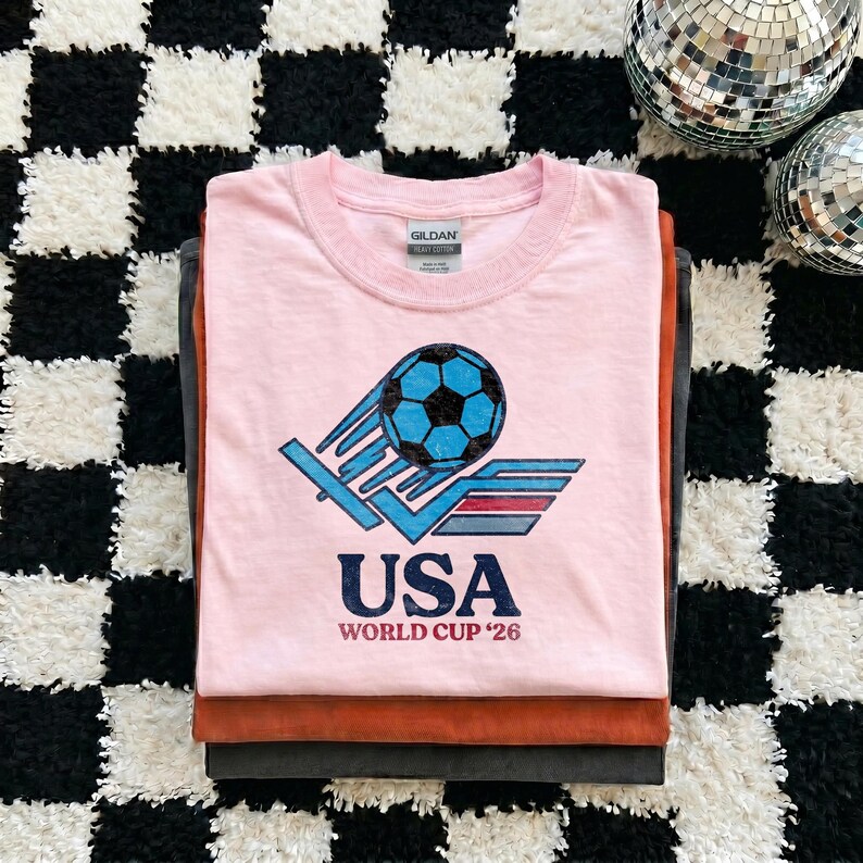 Puede incluir: Una pila de tres camisetas. La camiseta superior es de color rosa claro con un gr&aacute;fico de un bal&oacute;n de f&uacute;tbol y el texto "USA WORLD CUP '26". Las otras camisetas son marrones y grises.