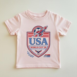 Peut inclure: Un t-shirt rose clair avec un graphisme pour la Coupe du Monde de la FIFA 2026 des &Eacute;tats-Unis. Le design comprend un ballon de football, une fus&eacute;e et le texte "USA" en bleu. Le t-shirt a des manches courtes et un col rond.