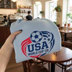 May include: A light blue sweatshirt featuring a soccer ball and the text "USA WORLD CUP '26" in red and blue. The design includes stars and a stylized flame element, promoting the upcoming World Cup.