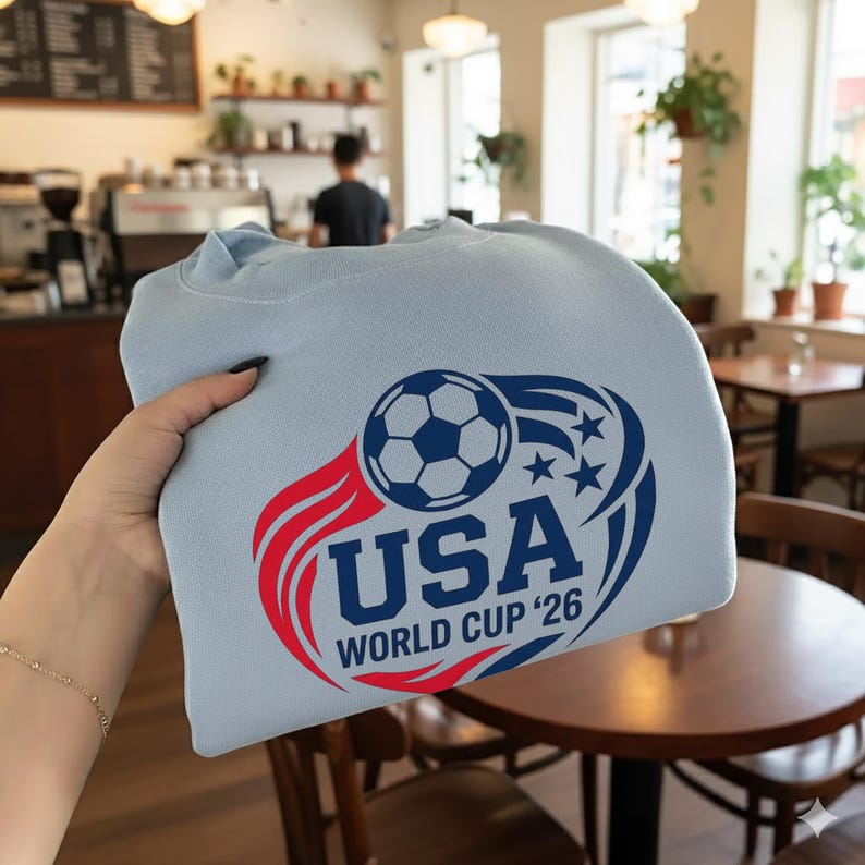 May include: A light blue sweatshirt featuring a soccer ball and the text "USA WORLD CUP '26" in red and blue. The design includes stars and a stylized flame element, promoting the upcoming World Cup.