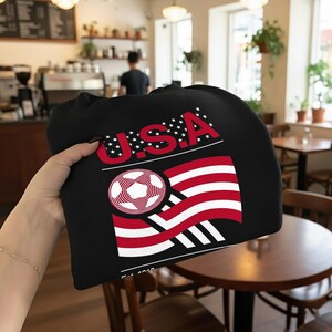 May include: Black sweatshirt featuring a graphic of the USA flag with a soccer ball design. The word "USA" is in red with white stars. The design includes red and white stripes and a soccer ball graphic.