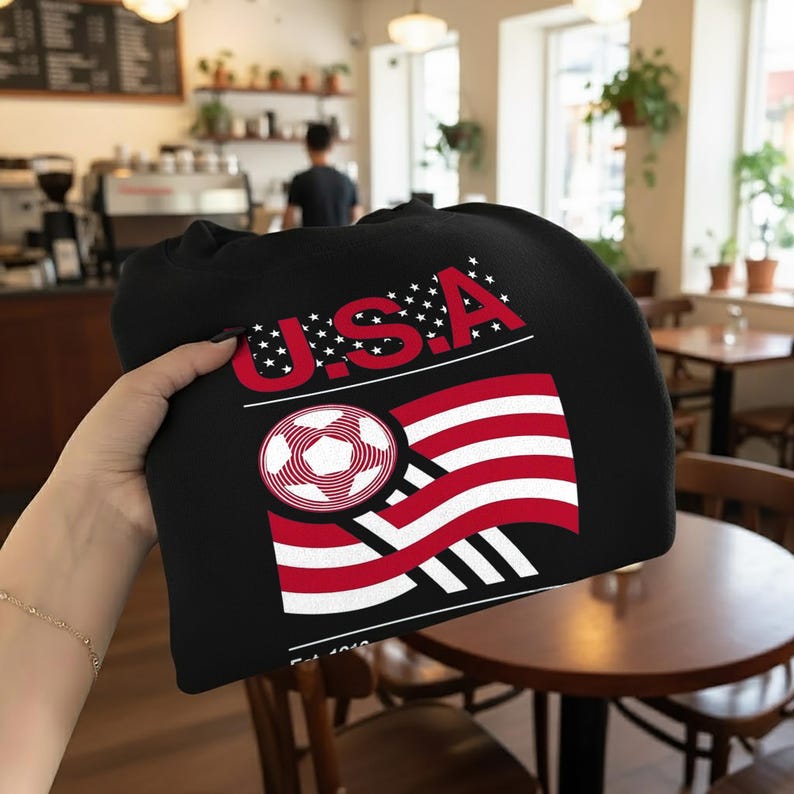 May include: Black sweatshirt featuring a graphic of the USA flag with a soccer ball design. The word "USA" is in red with white stars. The design includes red and white stripes and a soccer ball graphic.