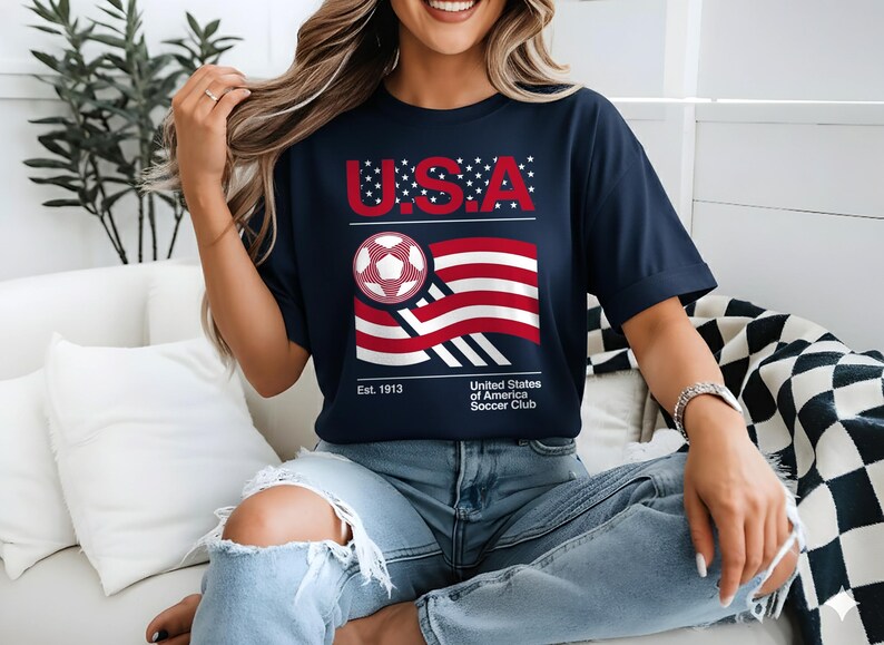 May include: Navy blue t-shirt with a graphic featuring the text "U.S.A." in red, a soccer ball, and a stylized American flag. The shirt also includes the text "Est. 1913" and "United States of America Soccer Club."