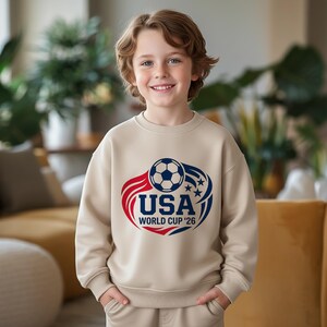 May include: A beige sweatshirt featuring a soccer ball graphic with the text "USA WORLD CUP '26" in red and blue. The design includes stars and curved lines. The sweatshirt is worn by a smiling child.