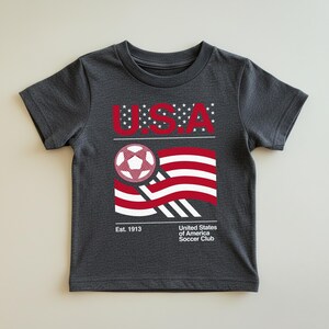 May include: Dark gray t-shirt featuring a graphic of a soccer ball and a stylized American flag in red and white. The word "USA" is above the flag, with the text "United States of America Soccer Club" and "Est. 1913" below.