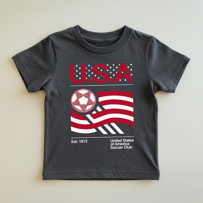 May include: Dark gray t-shirt featuring a graphic of a soccer ball and a stylized American flag in red and white. The word "USA" is above the flag, with the text "United States of America Soccer Club" and "Est. 1913" below.