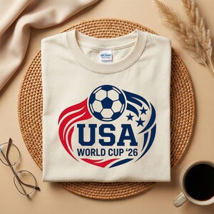 May include: Cream-colored t-shirt with a soccer ball graphic. The design features a soccer ball, the word "USA", and "World Cup '26" in red and blue. The shirt is on a woven mat with a coffee cup and glasses.