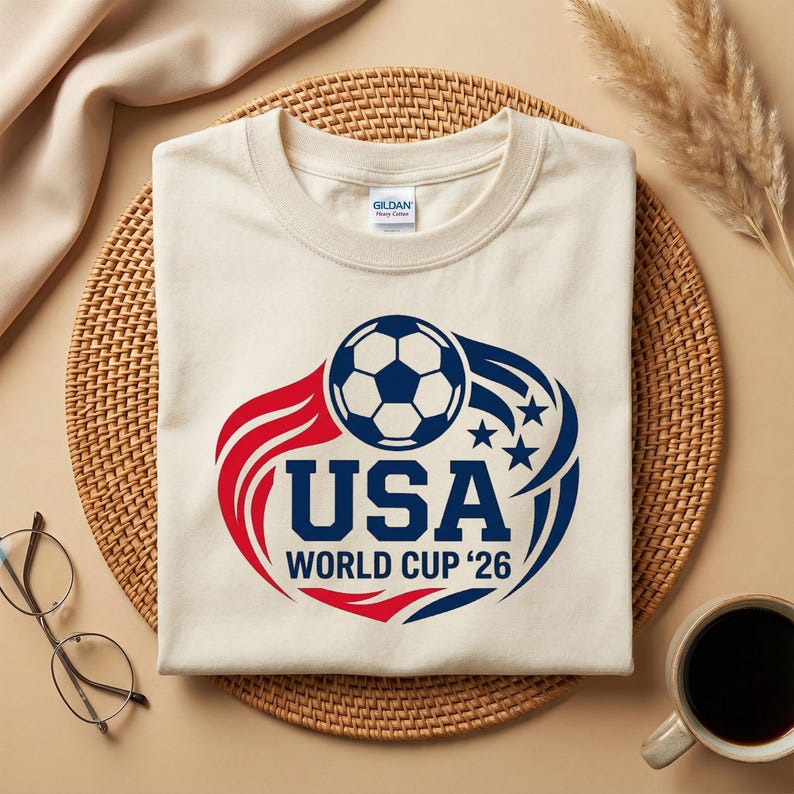 May include: Cream-colored t-shirt with a soccer ball graphic. The design features a soccer ball, the word "USA", and "World Cup '26" in red and blue. The shirt is on a woven mat with a coffee cup and glasses.