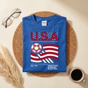 May include: Blue t-shirt with a graphic featuring "U.S.A." in red letters, a soccer ball, and a waving American flag design. The shirt also includes the text "Est. 1913" and "United States of America Soccer Club."