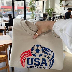 May include: Beige sweatshirt featuring a soccer ball graphic with red and blue accents, the word "USA", and "WORLD CUP '26". The design includes stars and flame-like elements, perfect for soccer fans.