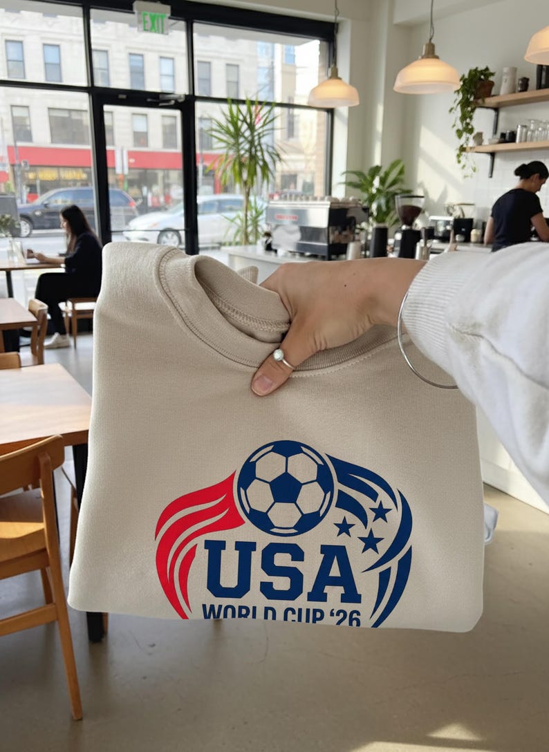 May include: Beige sweatshirt featuring a soccer ball graphic with red and blue accents, the word "USA", and "WORLD CUP '26". The design includes stars and flame-like elements, perfect for soccer fans.