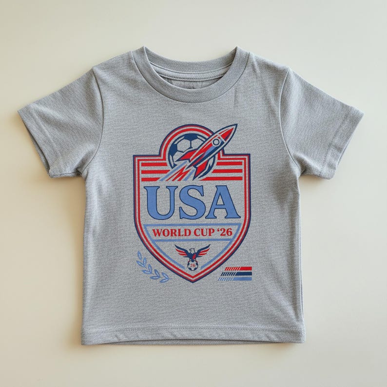 Peut inclure: Un t-shirt gris avec un motif de bouclier portant l'inscription "USA WORLD CUP '26". Le motif comprend un ballon de football, une fus&eacute;e et un aigle. Le t-shirt est &agrave; manches courtes.