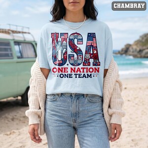 May include: A light blue t-shirt with the word "USA" in a patriotic design featuring the Statue of Liberty, American flags, and an eagle. The text "ONE NATION ONE TEAM" is below the graphic. The shirt is worn by a person on a beach.