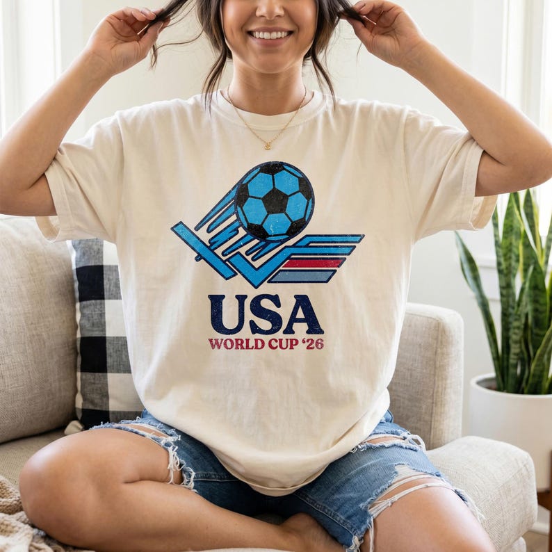 Puede incluir: Camiseta color crema con un bal&oacute;n de f&uacute;tbol azul y el texto "USA WORLD CUP '26". El dise&ntilde;o incluye l&iacute;neas de movimiento azules y un emblema rojo y azul en forma de ala. Aspecto vintage.