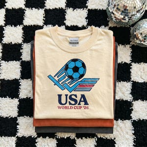 Puede incluir: Una pila de tres camisetas. La camiseta superior es de color crema con un gr&aacute;fico de un bal&oacute;n de f&uacute;tbol azul y el texto "USA WORLD CUP '26". Las camisetas est&aacute;n apiladas sobre una superficie a cuadros en blanco y negro.