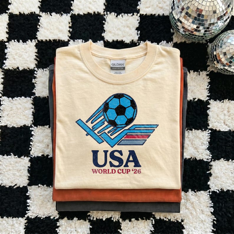 Puede incluir: Una pila de tres camisetas. La camiseta superior es de color crema con un gr&aacute;fico de un bal&oacute;n de f&uacute;tbol azul y el texto "USA WORLD CUP '26". Las camisetas est&aacute;n apiladas sobre una superficie a cuadros en blanco y negro.