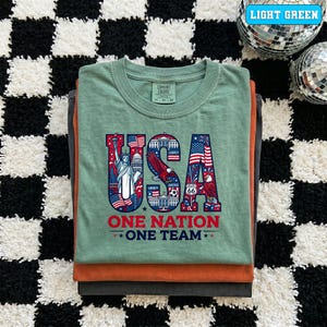 May include: A stack of folded t-shirts in shades of green, orange, and gray. The top shirt is light green and features a patriotic "USA" graphic with the Statue of Liberty, an eagle, and the text "ONE NATION ONE TEAM."
