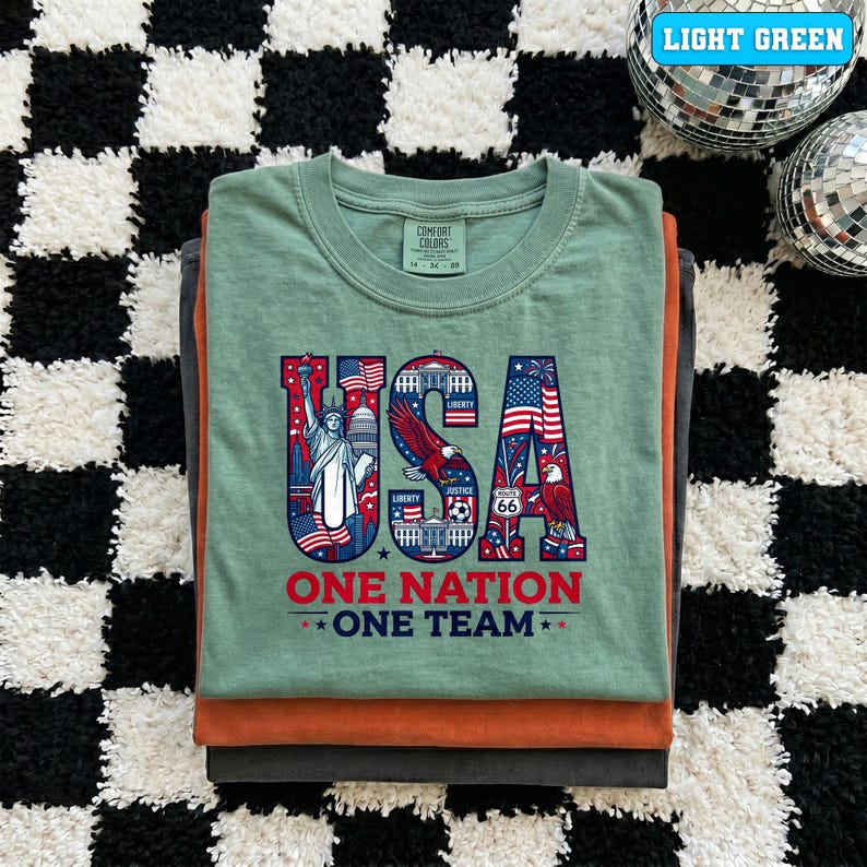 May include: A stack of folded t-shirts in shades of green, orange, and gray. The top shirt is light green and features a patriotic "USA" graphic with the Statue of Liberty, an eagle, and the text "ONE NATION ONE TEAM."
