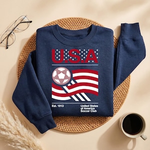May include: Navy blue sweatshirt with a graphic featuring the letters "U.S.A." above a soccer ball and a waving American flag design. The text "Est. 1913" and "United States of America Soccer Club" are also included.