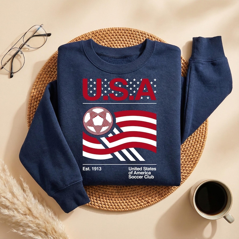 May include: Navy blue sweatshirt with a graphic featuring the letters "U.S.A." above a soccer ball and a waving American flag design. The text "Est. 1913" and "United States of America Soccer Club" are also included.