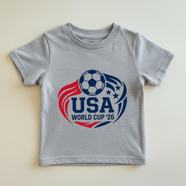 May include: A gray t-shirt featuring a soccer ball graphic with red and blue accents. The design includes the text "USA WORLD CUP '26" in navy blue, with stars and curved lines. The shirt is made of a soft material.