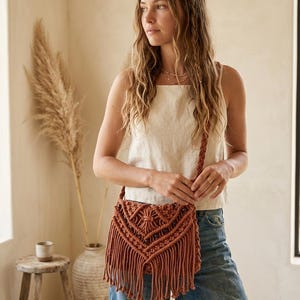 May include: A terracotta-colored macramé crossbody bag featuring an intricate knotted geometric pattern, a braided shoulder strap, and long fringe detailing along the bottom edge, accented with small dark wooden beads integrated into the design.