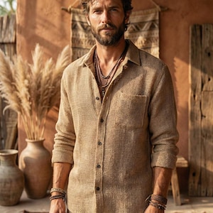 Puede incluir: Una camisa beige con botones y tejido texturizado, con bolsillo en el pecho. La camisa se combina con pantalones beige y collares superpuestos. Llevada con pulseras, el atuendo tiene un estilo relajado e informal.