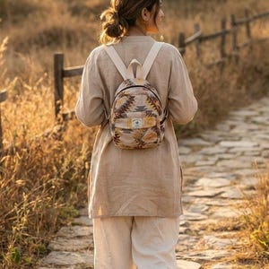 Puede incluir: Una mochila pequeña con un diseño estampado en tonos beige, marrón y amarillo. La mochila tiene correas color crema y una pequeña etiqueta. La persona lleva una túnica beige y pantalones blancos, caminando por un camino de piedra.