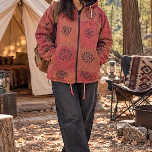 May include: A red and orange patchwork hooded jacket with black block-printed bird and floral motifs, a front zipper closure, and drawstring cords, paired with dark gray denim jeans, a brown knit beanie, and brown hiking boots.