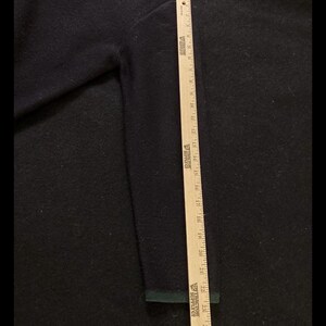 Puede incluir: Un su&eacute;ter azul marino con un ribete verde en el pu&ntilde;o. Se coloca una regla de madera junto a la manga para medir la longitud. El su&eacute;ter tiene un aspecto texturizado.