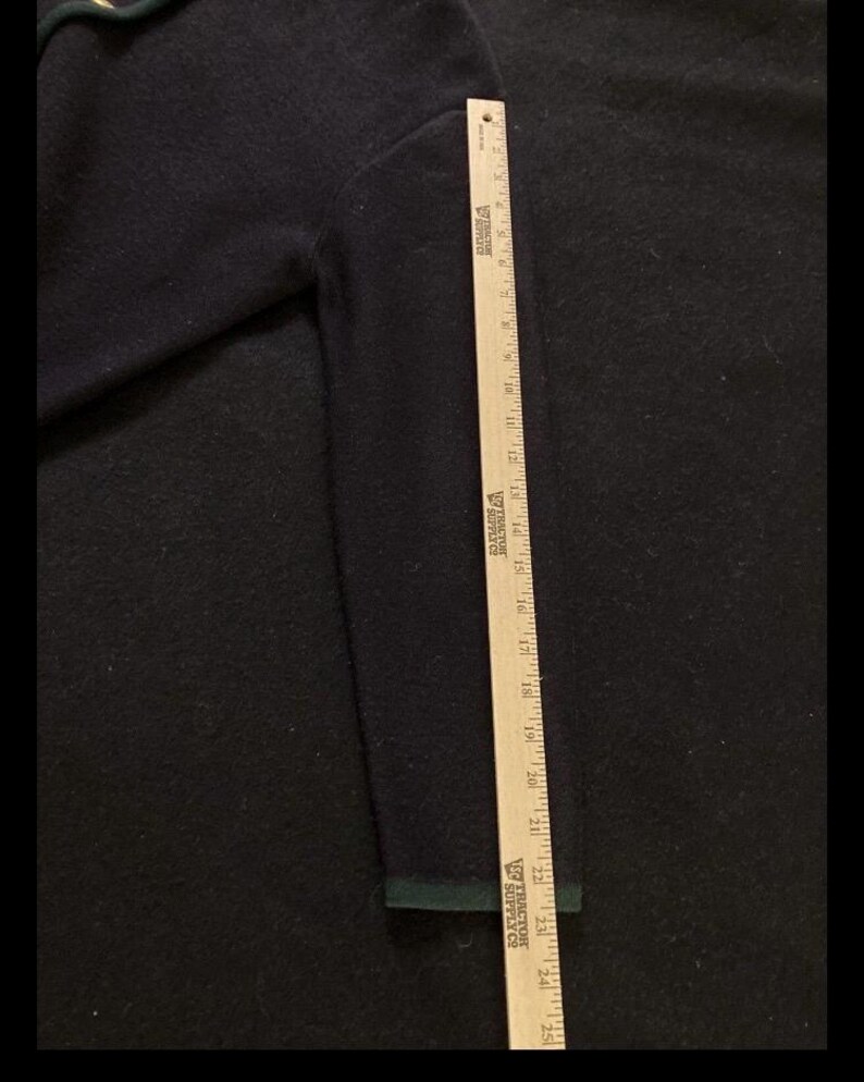 Puede incluir: Un su&eacute;ter azul marino con un ribete verde en el pu&ntilde;o. Se coloca una regla de madera junto a la manga para medir la longitud. El su&eacute;ter tiene un aspecto texturizado.