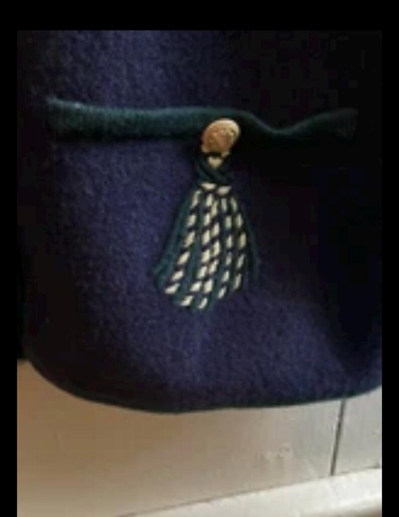 Puede incluir: Primer plano de una prenda azul marino con un ribete verde oscuro y una borla decorativa. La borla es blanca y verde oscuro, y la prenda tiene un bot&oacute;n dorado.