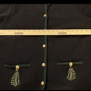 Puede incluir: Un c&aacute;rdigan azul marino oscuro con ribetes verdes y botones dorados. El c&aacute;rdigan tiene dos bolsillos con ribetes verdes y borlas decorativas. Una regla de madera se coloca sobre el c&aacute;rdigan, midiendo aproximadamente 51 cm.