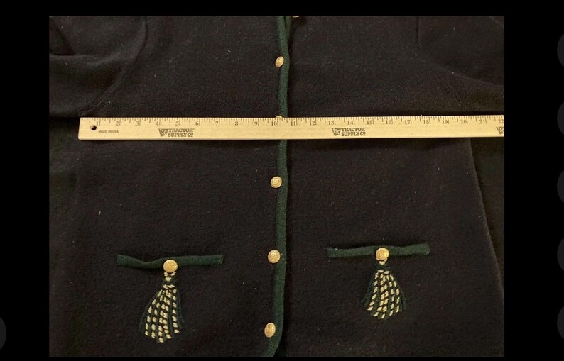 Puede incluir: Un c&aacute;rdigan azul marino oscuro con ribetes verdes y botones dorados. El c&aacute;rdigan tiene dos bolsillos con ribetes verdes y borlas decorativas. Una regla de madera se coloca sobre el c&aacute;rdigan, midiendo aproximadamente 51 cm.