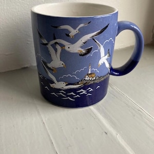 Op de afbeelding: Een blauwe keramische mok met een witte binnenkant en een gebogen handvat. De mok heeft een kustscène met vliegende zeemeeuwen boven een kustlijn met een vuurtoren. Het ontwerp is in blauw- en wittinten.