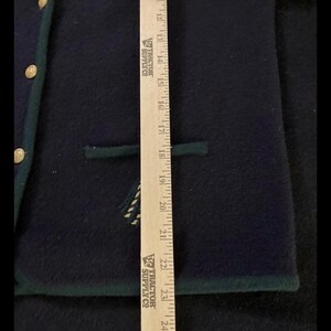 Puede incluir: Chaqueta de lana azul marino con ribete verde y botones dorados. La chaqueta tiene un bolsillo y un dise&ntilde;o decorativo. Una regla se coloca junto a la chaqueta, midiendo aproximadamente 61 cm.