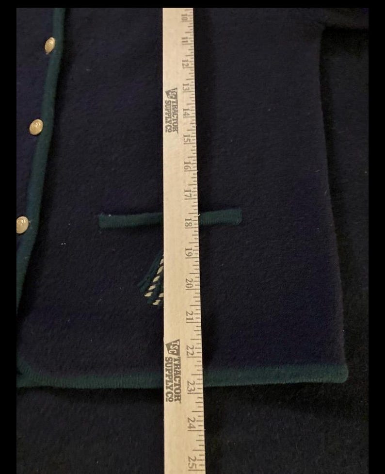 Puede incluir: Chaqueta de lana azul marino con ribete verde y botones dorados. La chaqueta tiene un bolsillo y un dise&ntilde;o decorativo. Una regla se coloca junto a la chaqueta, midiendo aproximadamente 61 cm.