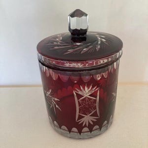 Op de afbeelding: Een robijnrode glazen pot met een deksel. De pot heeft ingewikkelde geëtste ontwerpen, waaronder sterren en bloempatronen. De deksel heeft een facetknop. De pot is waarschijnlijk voor opslag of display.